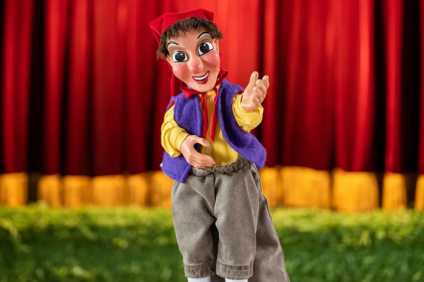 Der Kasperl aus Raphael Kovariks Kasperlkiste winkt den Kindern. Er steht auf einer grünen Wiese vor dem roten Vorhang.