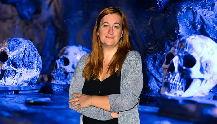 Ulrike Dziurzynski steht vor einem Escape Room, zu sehen sind Totenköpfe vor blauer Kulisse.