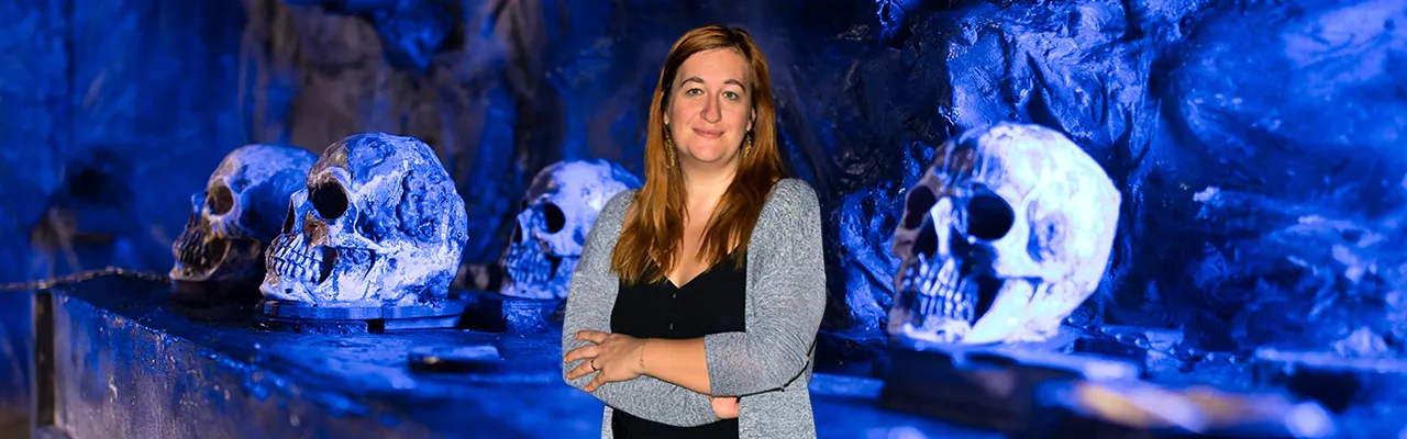 Ulrike Dziurzynski steht vor einem Escape Room, zu sehen sind Totenköpfe vor blauer Kulisse.